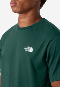 Camiseta de algodón verde oscuro con un logotipo blanco en el pecho izquierdo, cuello redondo y mangas cortas de corte clásico.