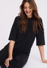 Młoda kobieta z długimi brązowymi włosami, ubrana w czarną, teksturowaną bluzkę i czarne spodnie, siedzi i uśmiecha się, spoglądając w dół.