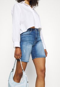 Camisa blanca corta de botones, pantalones cortos de mezclilla azules con detalles desgastados y un bolso azul claro con una correa.