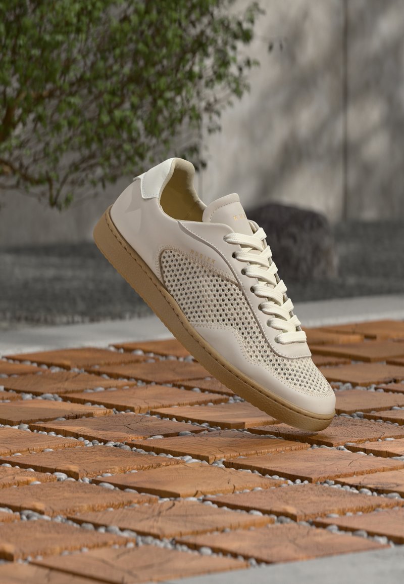 Baskets beige avec côtés en mesh et semelle en gomme flottant au-dessus d'une surface extérieure en bois texturé et pierre, avec un arrière-plan flou de verdure et de béton.