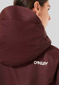 Maroon jacket with a high collar and large hood. Features white "Oakley" logo on the back. Smooth fabric with minimal texture.