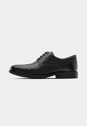 Zapato de vestir de piel negra con acabado brillante, punta redondeada, diseño con cordones, costuras contrastantes y un tacón bajo de goma.