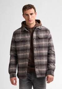Ruitjes shirtjack in bruin, grijs en crèmetinten, met twee borstzakken en drukknoopsluitingen, gedragen over een bruine hoodie.
