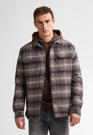 Veste à carreaux dans des tons marron, gris et crème, dotée de deux poches poitrine et de fermetures à boutons-pression, superposée à un hoodie marron.