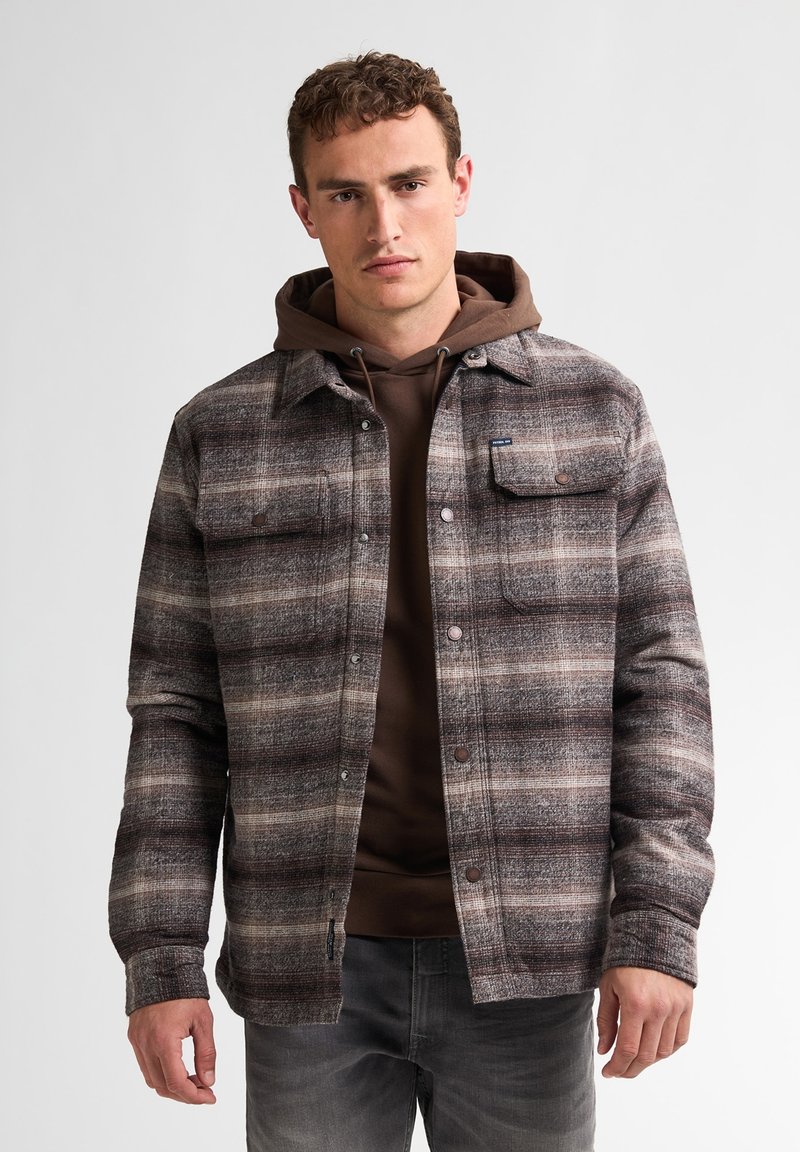 Ruitjes shirtjack in bruin, grijs en crèmetinten, met twee borstzakken en drukknoopsluitingen, gedragen over een bruine hoodie.
