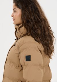 Kvinde med langt, krøllet hår iført en tan farvet pufferjakke med højt krave og sort logo-patch på ærmet, vendt mod venstre på en hvid baggrund.