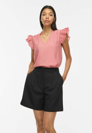 Femme aux longs cheveux foncés portant un chemisier rose à manches volantées et un short noir taille haute, debout les mains dans les poches sur fond blanc.