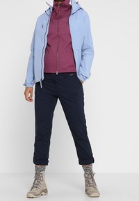 Světlomodrá lehká bunda přes bordó fleecovou bundu na zip; kombinováno s tmavě modrými kalhotami a béžovými turistickými botami, doplněno pletenými šedými ponožkami.