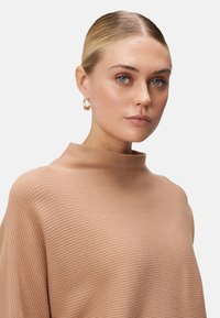 Jeune femme avec des cheveux blonds gominés, portant des boucles d'oreilles en or et un pull col roulé beige côtelé, posant devant un fond clair.