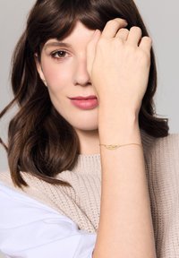 Złoty łańcuszek bransoletki z motywem liścia, z misternymi detalami. Modelka ma na sobie lekki sweter dzianinowy i eksponuje swoją rękę.