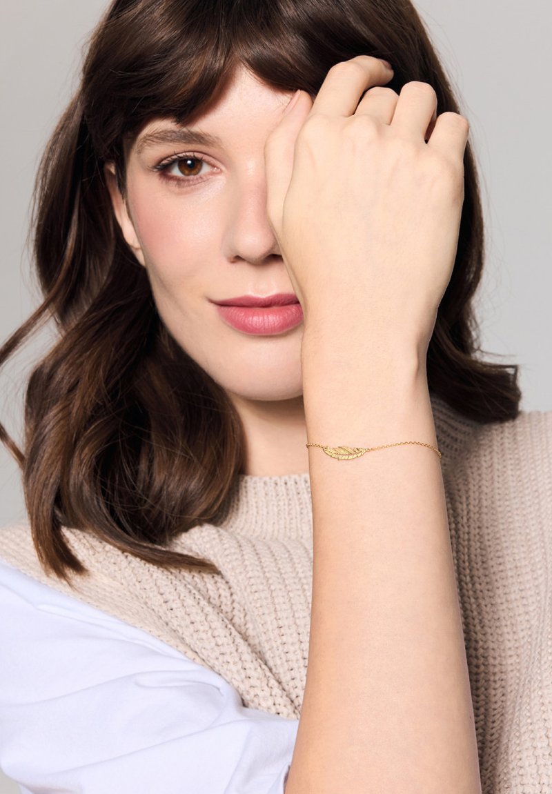 Złoty łańcuszek bransoletki z motywem liścia, z misternymi detalami. Modelka ma na sobie lekki sweter dzianinowy i eksponuje swoją rękę.