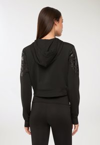 Donna con lunghi capelli castani che indossa un hoodie corto nero con toppe decorate sulle spalle e leggings neri, rivolta di spalle.
