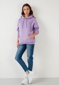 Sudadera con capucha lavanda con un bolsillo frontal, combinada con jeans skinny azules con un desgarro y zapatillas blancas gruesas con calcetines de rayas coloridas.
