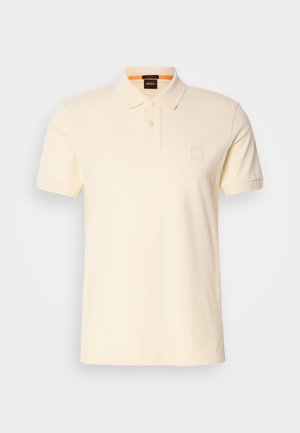 Polo em algodão bege claro com colarinho clássico, mangas curtas e um pequeno logotipo no peito. Textura suave, design minimalista.