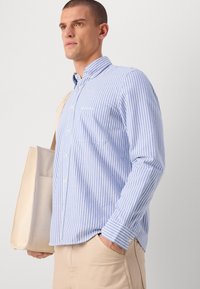 Blå og hvitstripete skjorte med krage, enkelt brystlomme og lange ermer. Materialet ser lett ut, kombinert med beige bukser.