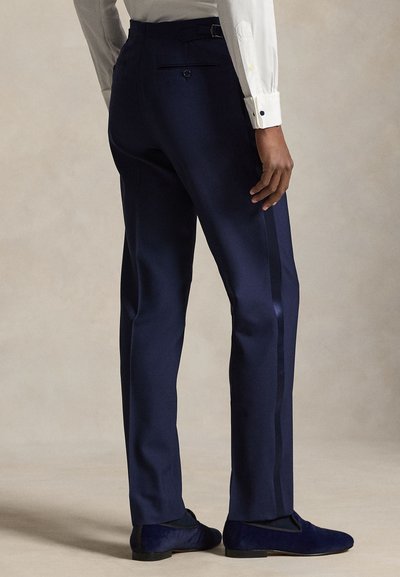 Άνδρας που φοράει ναυτικές παντελόνες με ρίγα στο πλάι και σκούρα μπλε μοκασίνια, στέκεται μπροστά σε απλό μπεζ φόντο.