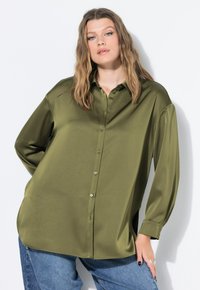 Giovane donna con capelli mossi che indossa una camicia oversize di satin verde oliva e jeans blue, posando contro uno sfondo bianco.