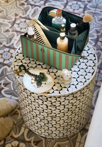 Round, mosaic table with black and white geometric pattern. A striped cosmetic bag on top, containing various bottles and a comb.