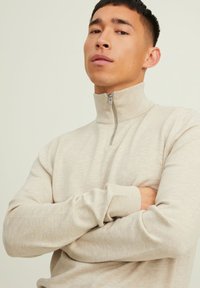 Suéter beige con cremallera y cuello alto, hecho de un tejido de punto texturizado. Presenta puños y dobladillo acanalados, con un diseño de corte ajustado.
