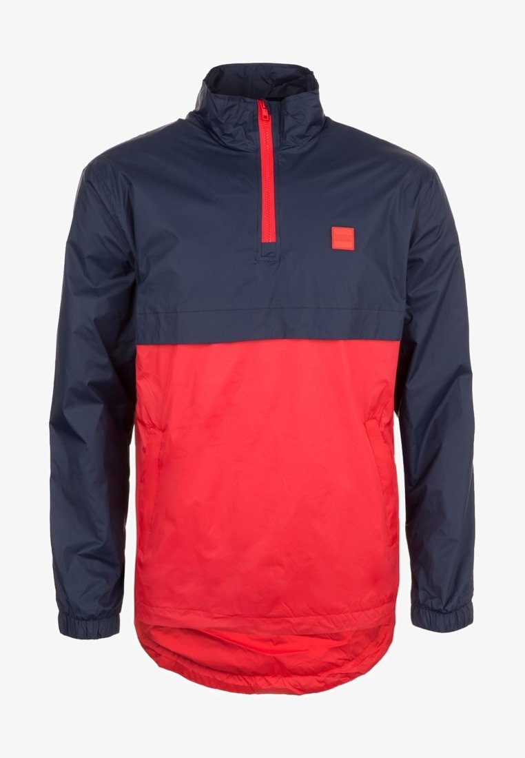 Windbreaker jacket featuring a half-zip design, navy blue upper section, bright red lower section, and elastic cuffs with a square logo patch.