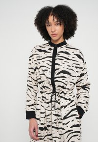 Šaty s dlhým rukávom z béžovej látky s čiernymi tigrovými pruhmi, s golierom na gombíky, sťahovacím pásom a čiernymi manžetami.