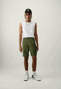 Green cargo shorts with multiple pockets, white sleeveless shirt, white sneakers, black cap, and sunglasses. Simple textures and casual design.