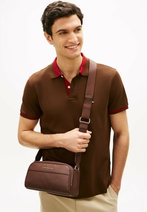 Jonge man die glimlacht, gekleed in een bruine poloshirt met een rode kraag en een bruine crossbodytas draagt over beige broeken.