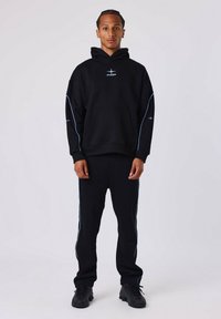 Jonge man met gevlochten haar, gekleed in een zwarte hoodie en bijpassende trainingsbroek met lichtblauwe piping, staand voor een plain witte achtergrond.