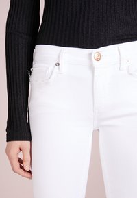 Jeans blancos ajustados con bordes desflecados en los bolsillos y cierre de botón dorado, combinados con una blusa de manga larga negra ajustada de canalé.