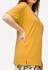 Donna che indossa un top a maniche corte giallo senape e pantaloni con fantasia, con la mano appoggiata sull'anca, il torso visibile di profilo.