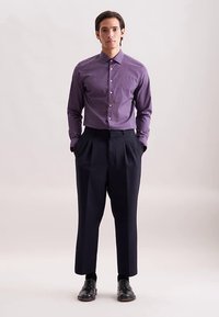 Camicia viola con bottoni e pois bianchi, abbinata a pantaloni scuri a vita alta e scarpe di pelle nere. Tessuto liscio, vestibilità sartoriale.