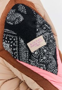 Hnědá, růžová a černá prošívaná bunda s částí z černé látky se vzorem a růžovým štítkem s inspirativním citátem.