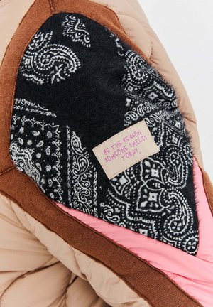 Część brązowej, różowej i czarnej pikowanej kurtki z czarną łatą z wzorem, na której znajduje się różowa etykieta z inspirującym cytatem.