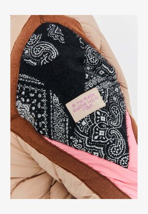 Część brązowej, różowej i czarnej pikowanej kurtki z czarną łatą z wzorem, na której znajduje się różowa etykieta z inspirującym cytatem.