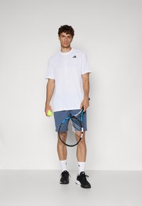 Weißes sportliches Shirt mit kurzen Ärmeln und dezentem Muster, kombiniert mit blauen Shorts. Hält einen Tennisschläger und einen Ball, trägt schwarze Sneakers.