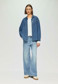 Een denimjack in medium blauw met een relaxte pasvorm, knoopsluiting en twee borstzakken. Draagt over een wit gebreid topje en lichtblauwe wijde jeans.