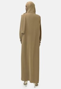 Langes, braunes Kleid mit langen Ärmeln und passendem Hijab. Glattes, leichtes Material und raffinierten Details an den Ärmeln, kombiniert mit hohen Absätzen.