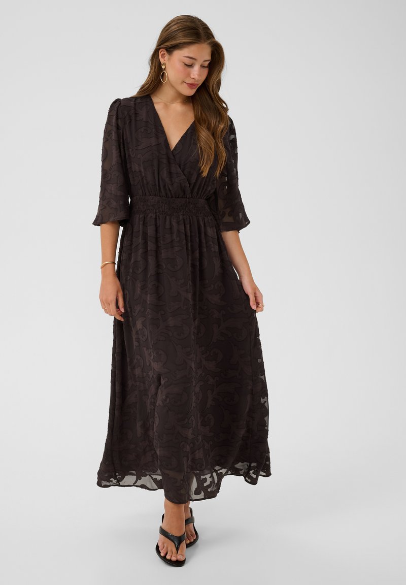 Femme aux cheveux longs portant une robe midi sombre à motifs avec des manches trois-quarts et des sandales noires, debout sur un fond clair uni.