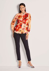 Frau steht da in einer Pfirsichbluse mit rot-blumigem Muster, schwarzen schmalen Hosen und mehrfarbigen High-Heels vor einem schlichten Hintergrund.