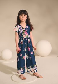 Temno modra cvetna kombinezon z kratkimi rokavi, elastičnim pasom in širokimi hlačnicami. Okrašena je z rožnatimi, vijoličnimi in belimi cvetovi. Oblečena z lahkimi sandali.