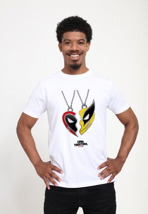 Man smiling with hands on hips wearing a white shirt showing two heart-shaped pendants with words "Best" and "Friends."