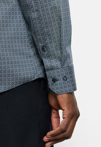 Marineblaues Shirt mit weißem geometrischem Muster, lange Ärmel, Knopfmanschetten mit zwei schwarzen Knöpfen, aus leichtem Stoff gefertigt.