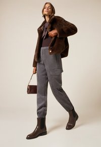 Chaqueta de piel sintética marrón, blusa oscura con lazo, pantalones gris rayados con bolsillos tipo cargo y botines marrones. Bolsa pequeña marrón en la mano.