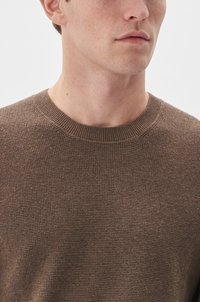 Braun strukturierter Strickpullover mit einem gerippten Rundhalsausschnitt, der eine Nahaufnahme des Stoffes sowie einen Teil des Halses und des Kinns des Models zeigt.