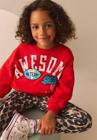 Sweatshirt rouge avec le texte "AWESOME" et le logo de l'équipe, associé à un legging à imprimé léopard. Des baskets montantes blanches complètent la tenue.