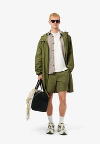 Olivgrön vattentät jacka, ljusgrå skjorta, vit T-shirt, gröna shorts, svart väska, mönstrad scarf, grå strumpor och sneakers.