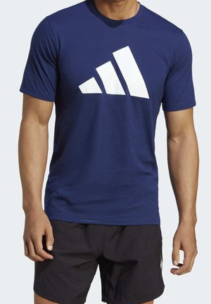 Un modèle cadré partiellement porte un T-shirt Adidas bleu marine avec un grand logo blanc et un short noir sur un fond clair.
