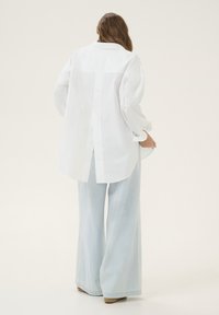 Woman wearing a loose white long-sleeve shirt with a back slit and light blue wide-leg jeans, standing facing away on a plain background.