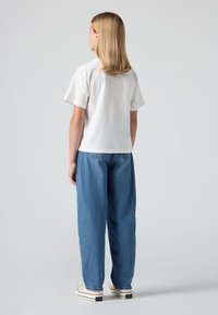 Mlada oseba z lasmi do ramen, obrnjena proč, oblečena v belo kratko majico z kratkimi rokavi, modre kavbojke in bele superge.