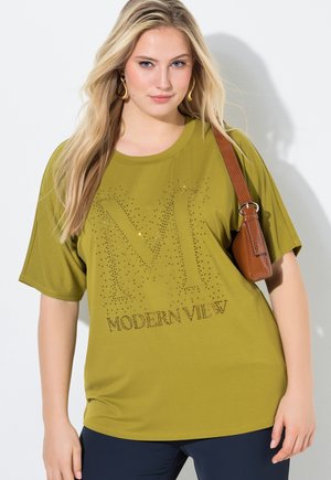 Lyshåret kvinde iført olivengrøn T-shirt med rhinsten "M Modern View"-design, marineblå bukser og en brun skuldertaske.